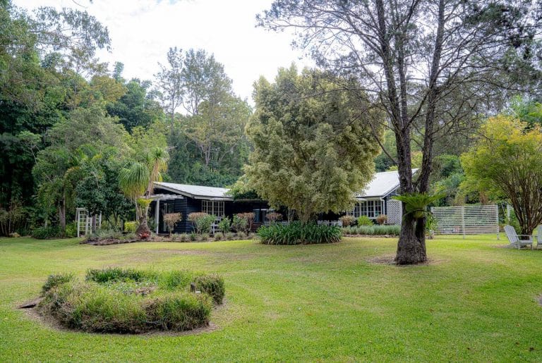Bahche House, a dark timber home with white trim, seen from its lawn under big trees