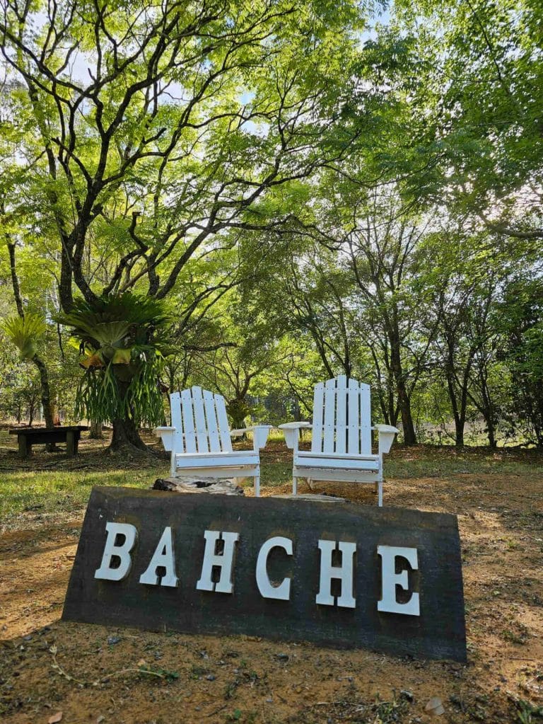 Two white chairs under the trees beside the Bahche sign